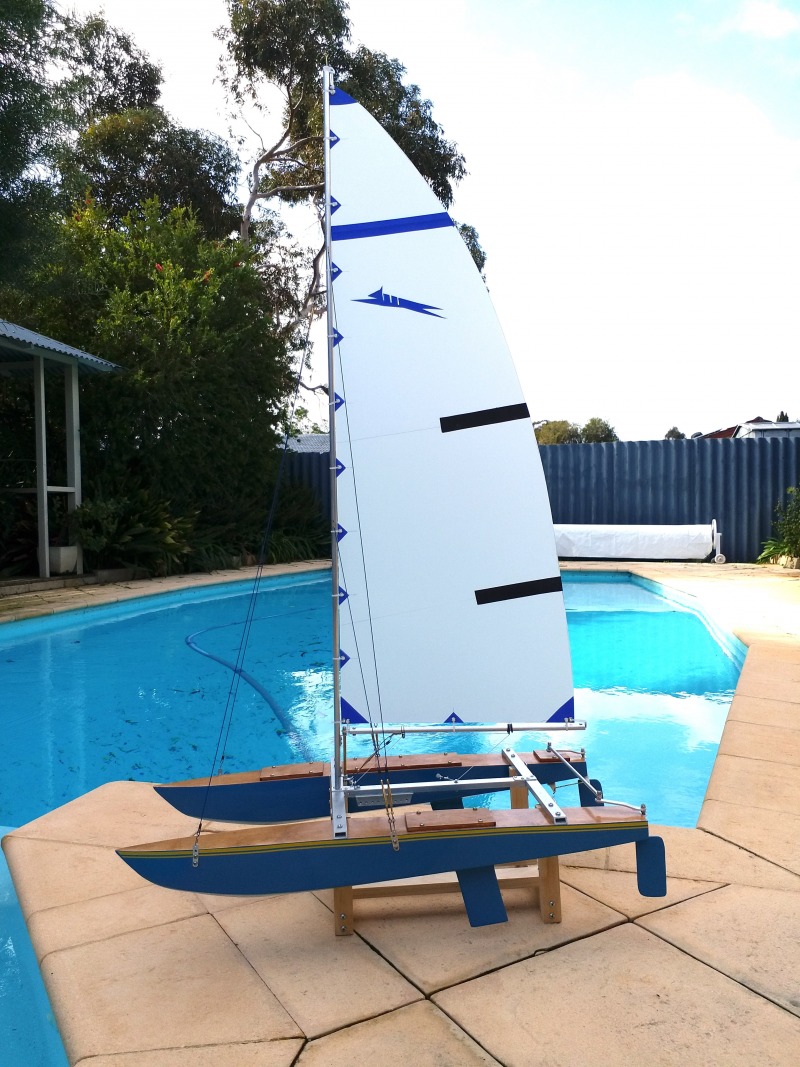 A radio controlled model yacht of a paper tiger catamaran. The hulls are both blue and it looks very much to scale of the real dinghy sailboat. It is sitting on a cradle next to a swimming pool, Location: Perth, Western Australia
