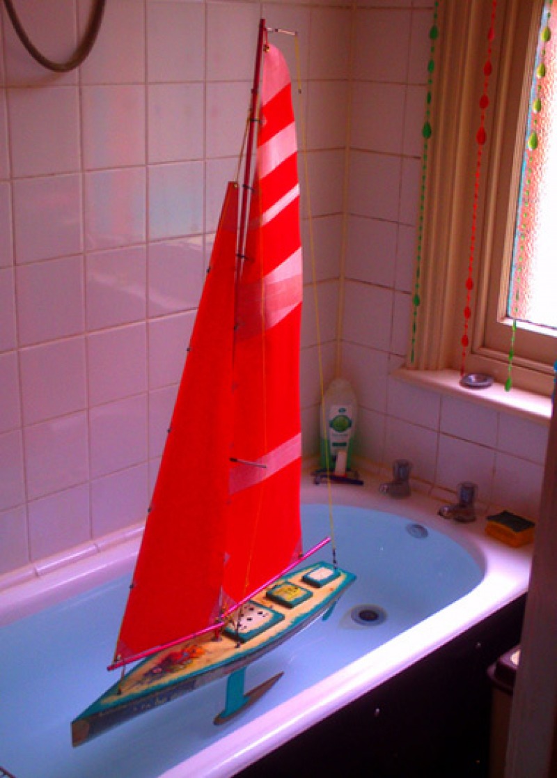 A colourful model yacht with red sails and bright blue swirly painted hull, sitting in a bathtub for it's first "float", Location: Portsmouth, England