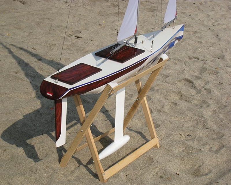 A model yacht sitting on a cradle on the beach, golden sand. The boat has lovely blue wavy graphics at the bow and faux-wood at the back in a swirly transition, Location: Indonesia