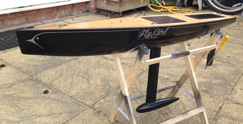 A model yacht sitting in a cradle without rigging, a black hull with swirly writing that says "Fly Girl"