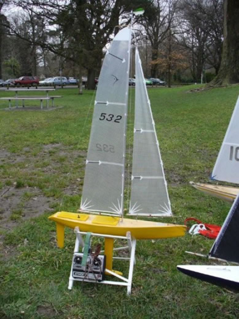 A yellow model yacht sitting in a cradle on a green field with controller leaning up against the cradle, Location: Christchurch, New Zealand
