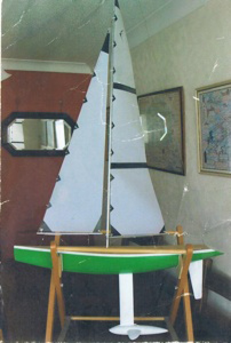 A large green rc yacht with a finned lead keel sits in a cradle in the living room. The sails are almost touching the ceiling it is so tall, Location: Internet